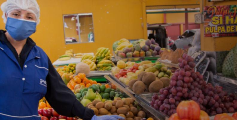 Mujer con indumentaria para manipulación de alimentos al lado de una plaza de mercado con frutas y verduras