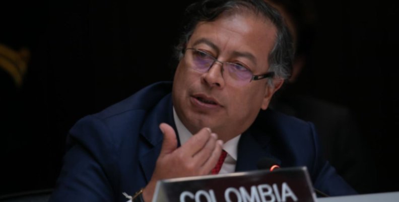 Petro Hombre con traje negro, gafas y corbata roja. Está sentado frente a una placa que dice "Colombia"