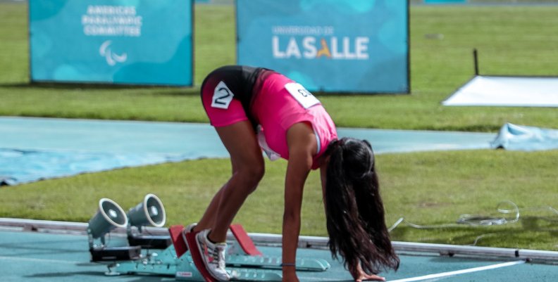 Juegos Parapanamericanos Atleta vestida de rosado en posición de salida. Está encima de un partidor de metal y una pista atlética azul