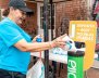 Mujer con camiseta azul depositando una botella en un cesto verde 