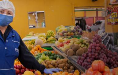 Mujer con indumentaria para manipulación de alimentos al lado de una plaza de mercado con frutas y verduras