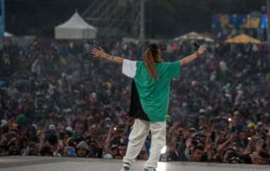 Hombre en el escenario con camisa verde y pantalón blanco abre los brazos frente al público